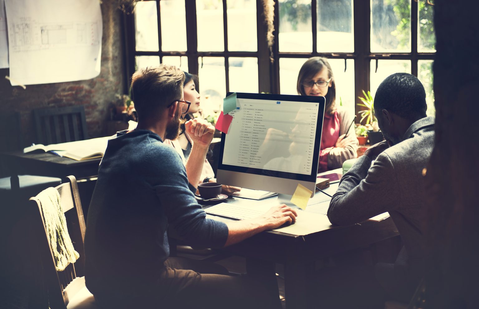 A group of diverse professionals collaborating around a desk in a modern office, with one person working on a desktop computer and others engaged in discussion during a team meeting.
