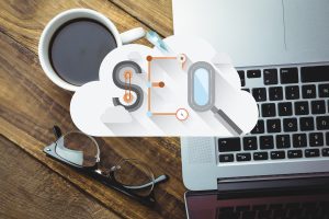 Top view of a workspace with a laptop, coffee cup, glasses, and an SEO cloud graphic featuring search and optimization icons.