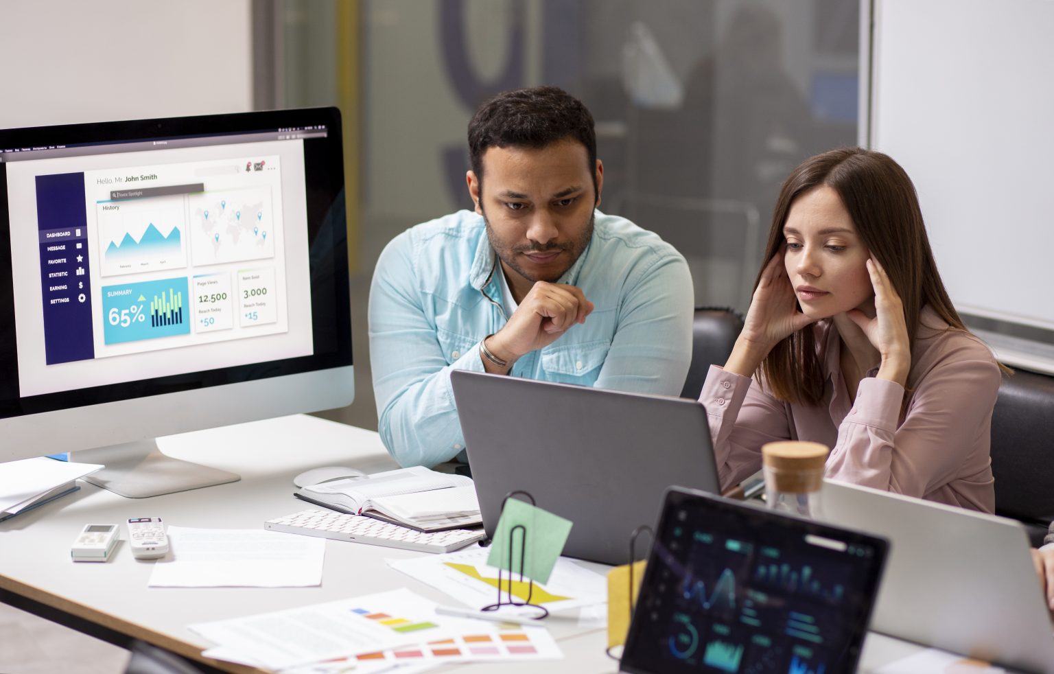 Two professional designers, a man and a woman, working intently in a modern office or computer lab. They are focused on a laptop screen while a large desktop monitor nearby displays a colorful data analytics dashboard with graphs and charts. The desk is organized with notebooks, tablets showing additional data, and design reference materials.