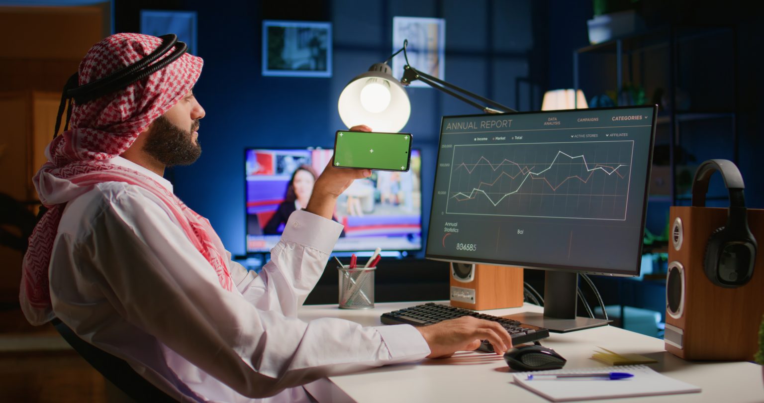 An Arab businessman tracking performance metrics on a computer and holding a green screen smartphone, showing the data analytics services of a digital marketing agency Dubai.