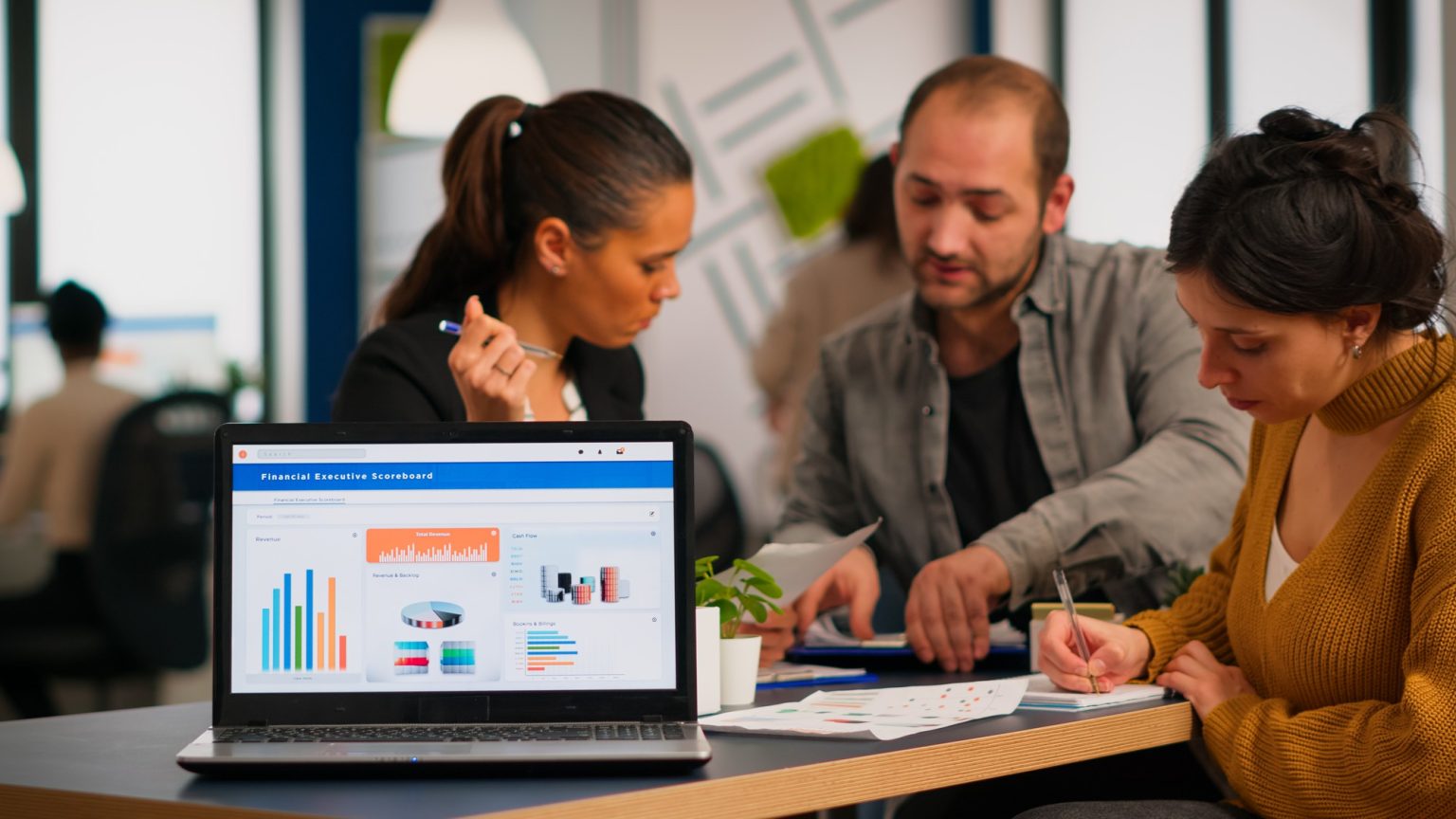 A digital marketing and financial analytics dashboard open on a laptop during a corporate team meeting in a busy agency workspace.