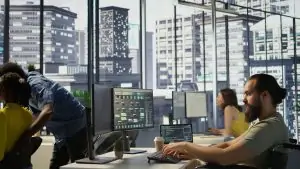 An inclusive team of developers working in a modern, high-rise office with large windows overlooking a city skyline. In the foreground, a man with a beard sits in a wheelchair at a desk, focused on coding on a dual-monitor setup. In the background, other colleagues are engaged at their workstations, creating a collaborative professional atmosphere.