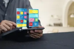 A person holding a tablet computer that projects a holographic smart home automation interface with colorful icons for CCTV, lighting, temperature, and security system controls.