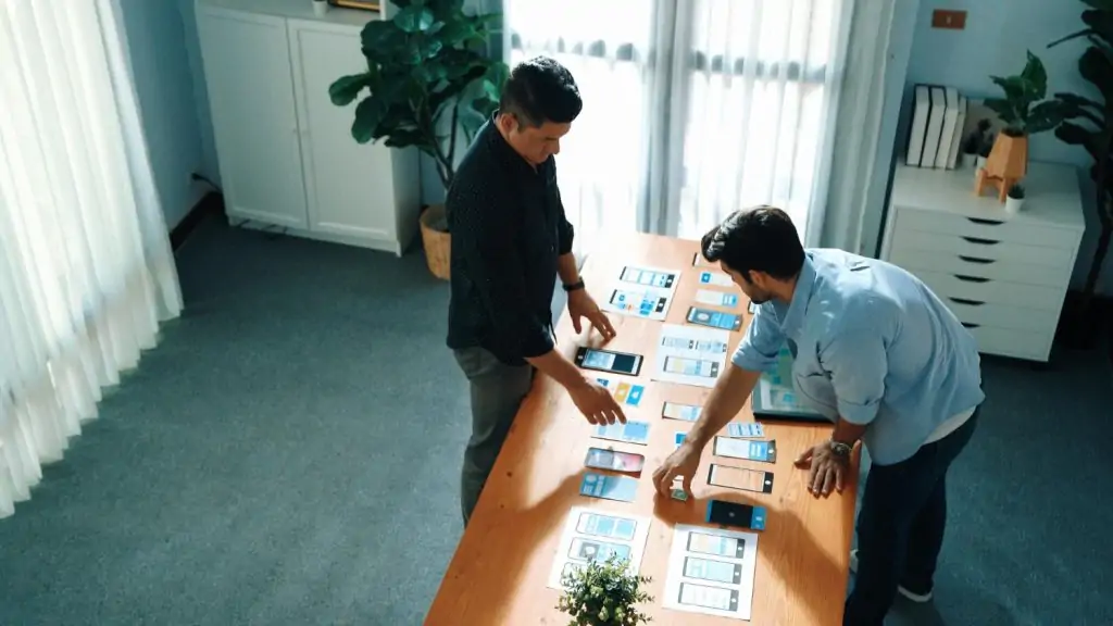 Two app developers standing at a wooden table reviewing printed mobile UI app interface wireframes and designs.